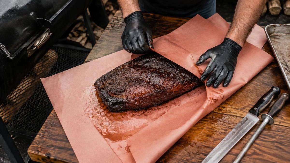 A brisket being wrapped in pink butcher paper on a cutting board
